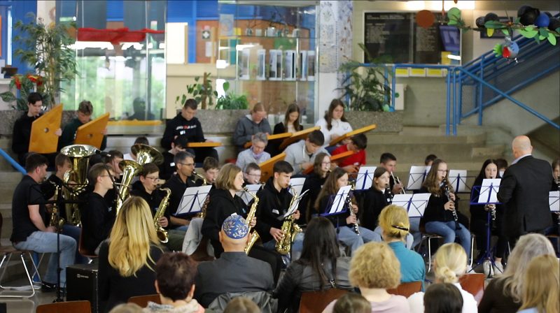 Blick auf das Schulorchester mit Blech- und Holzbläsern, Zauberharfen, Dirigent und Publikum