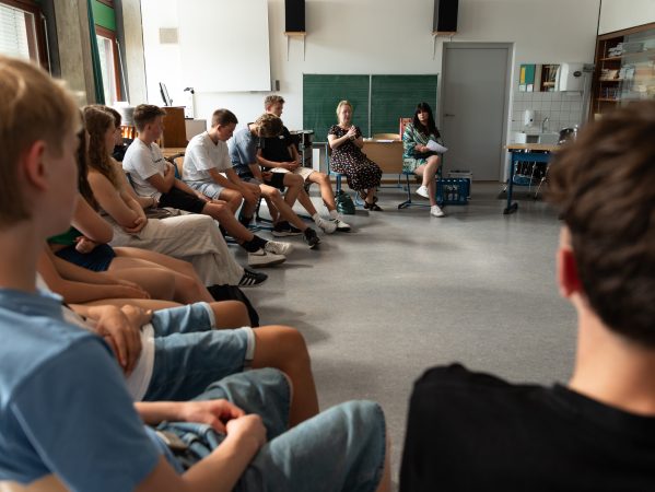 Yael Deusel Fiona Atay-Sandyk sprechen in einem Klassenzimmer im Kreis von Schülerinnen und Schülern